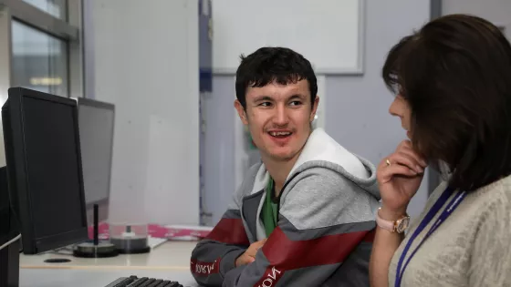 Male student sitting at desk with computer, talking to a female educator